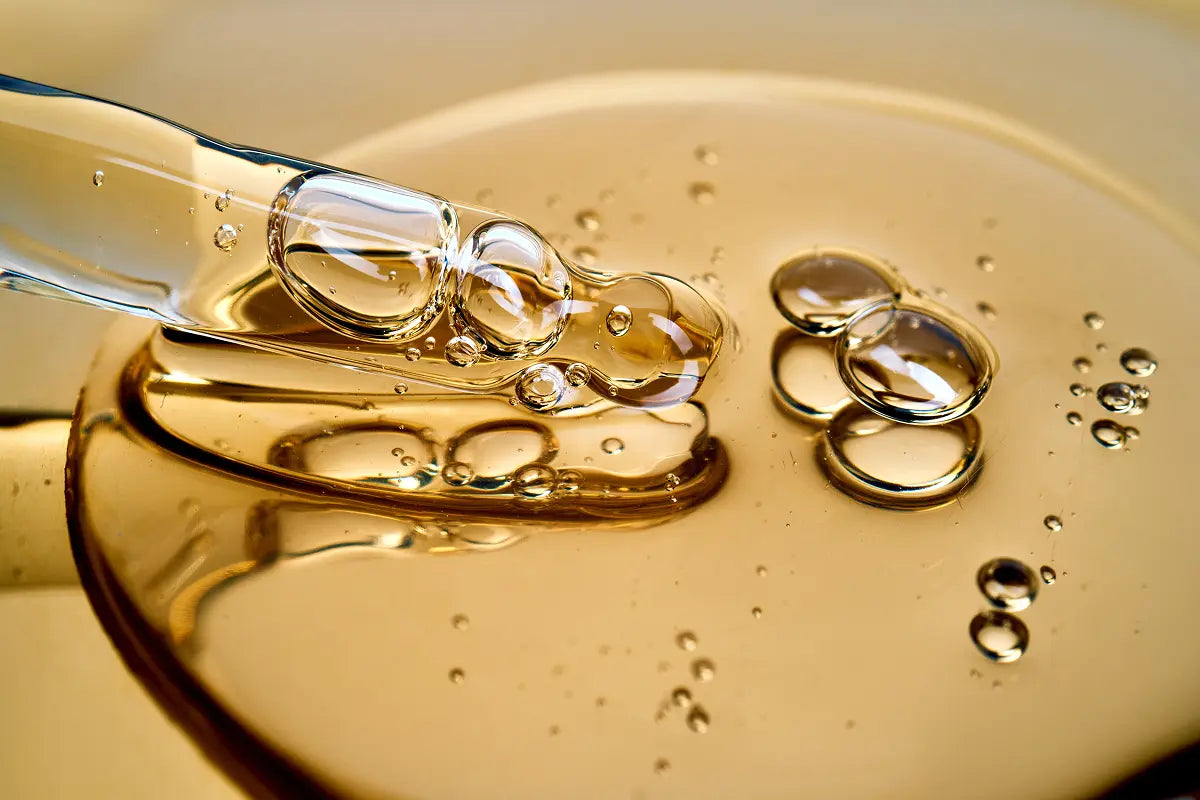 Macro view of clear serum being dispensed from a glass pipette into a golden gel, with fine bubbles suspended throughout