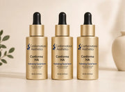 Three gold dropper bottles of Conformational Aesthetics Conforma HA Hydrating Facial Serum arranged side by side on a light beige surface with a soft neutral background.