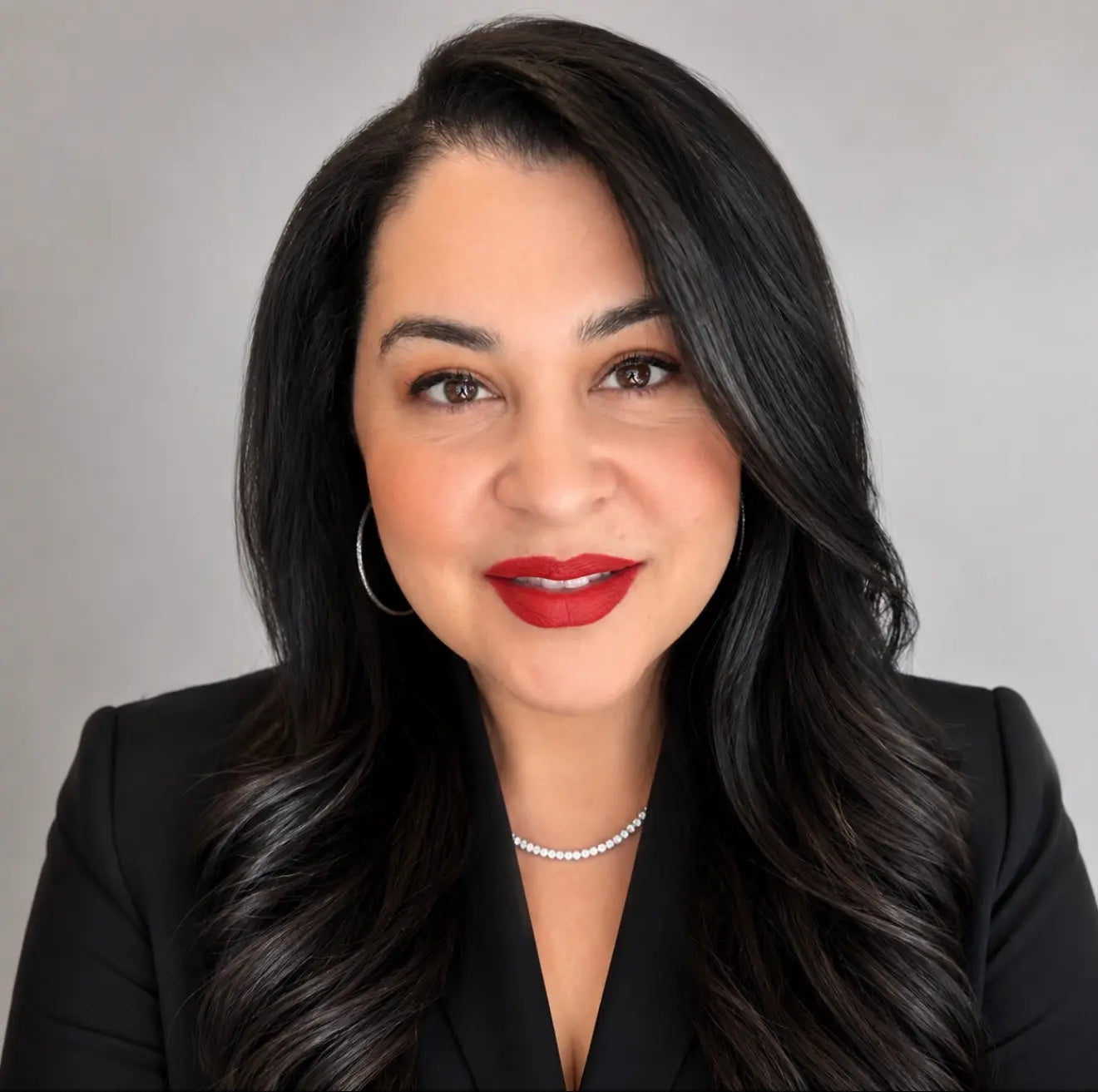 Portrait of Angelina Silva, Chief Operating Officer of Conformational Aesthetics. wearing a black blazer, red lipstick, hoop earrings, and a silver necklace against a light gray background.