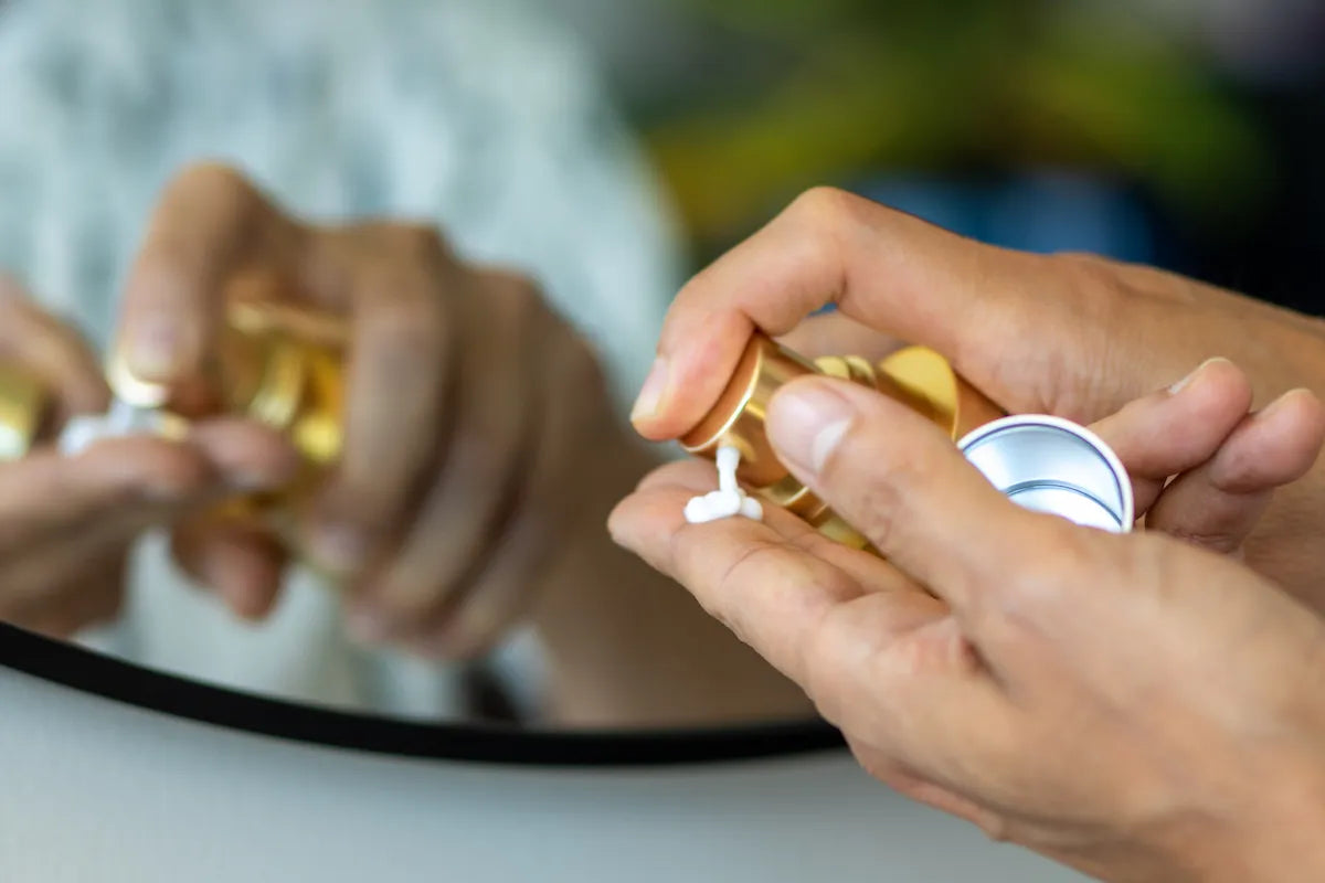 Hands dispensing Conforma Reduce cream from gold pump bottle in front of mirror