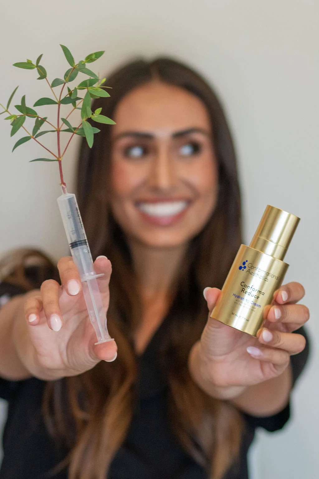 Smiling skincare professional holding a syringe with a leafy plant cutting and a gold bottle of Conforma Reducer cream.